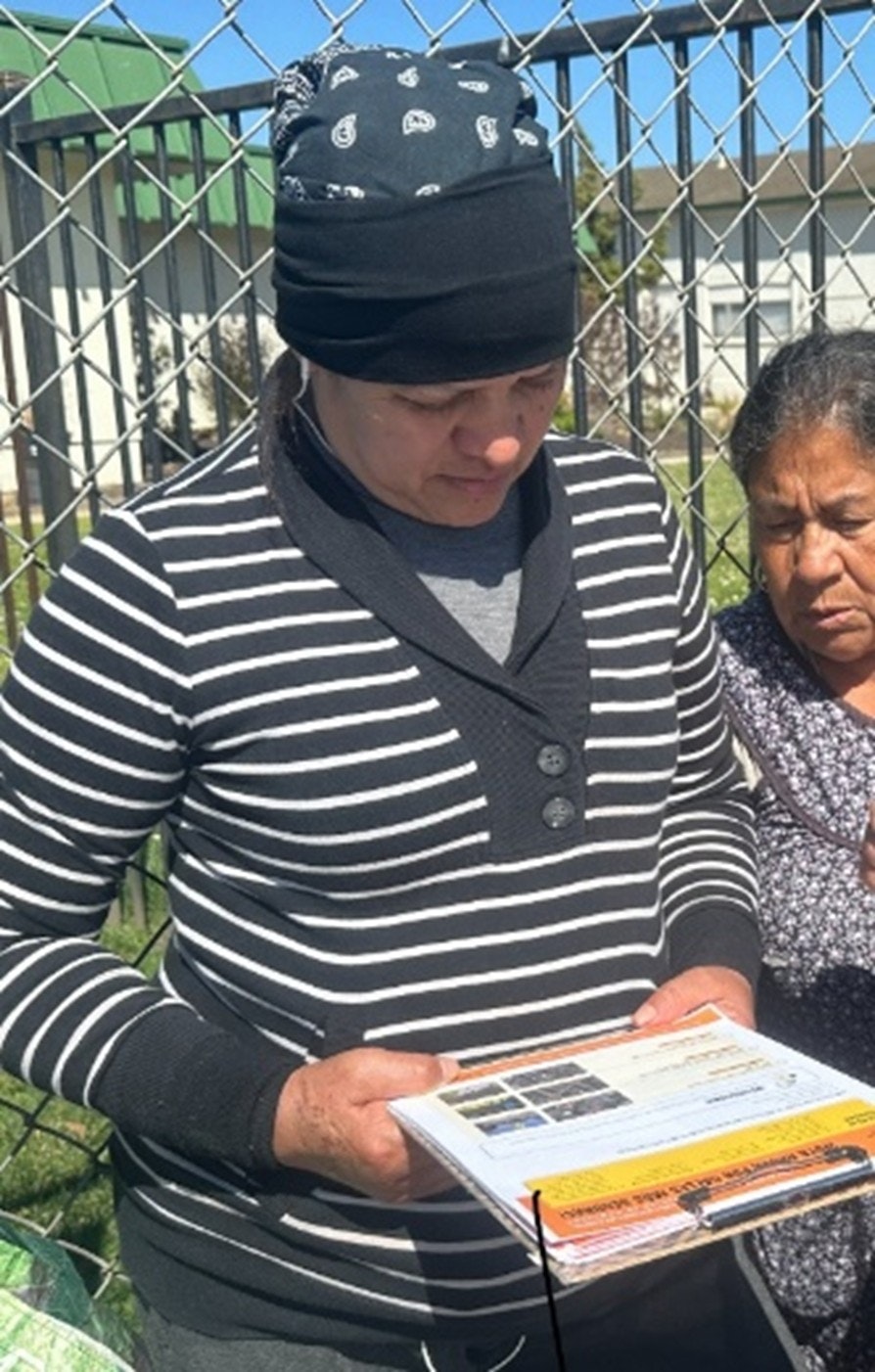 Two people stand in front of a fence. One is holding a clipboard with papers.