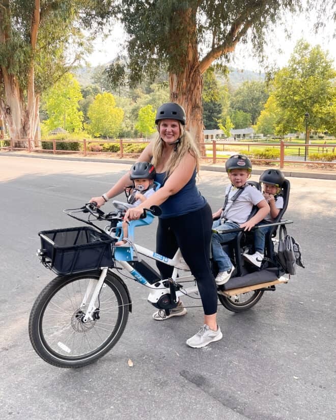 Woman riding a bike with three children seated, all wearing helmets, in a sunny park setting.