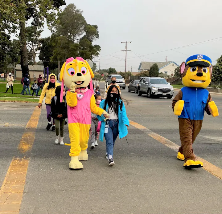 A group of children and adults in masks crossing the street, accompanied by costumed characters resembling cartoon dogs.