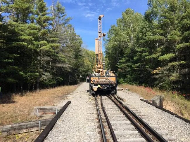 A rail-mounted machine on train tracks surrounded by trees under a clear sky.
