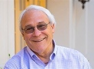 The image shows an older man smiling and wearing glasses, dressed in a light blue checkered shirt, with a pleasant background.