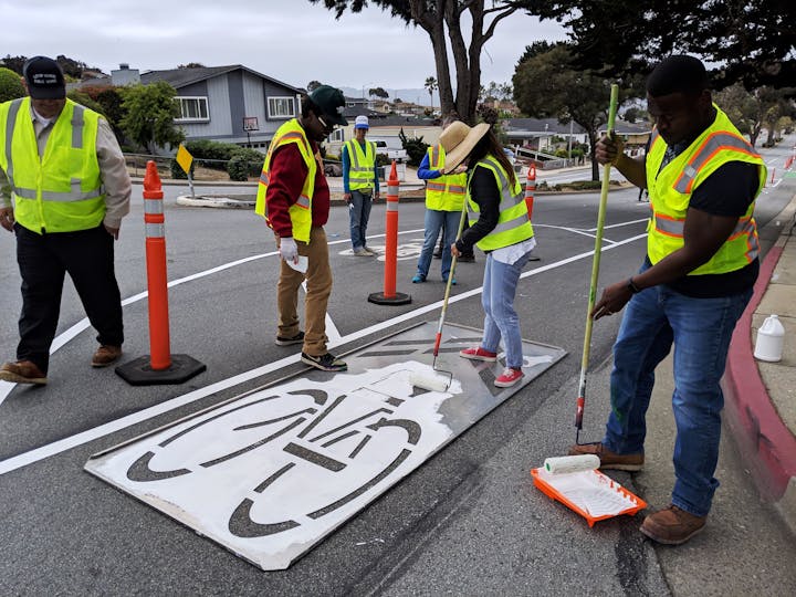 People in safety vests painting a bicycle lane symbol on the road with stencils.