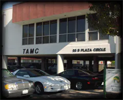The image shows a building with the acronym "TAMC" and an address at 55 B Plaza Circle, along with parked cars nearby.