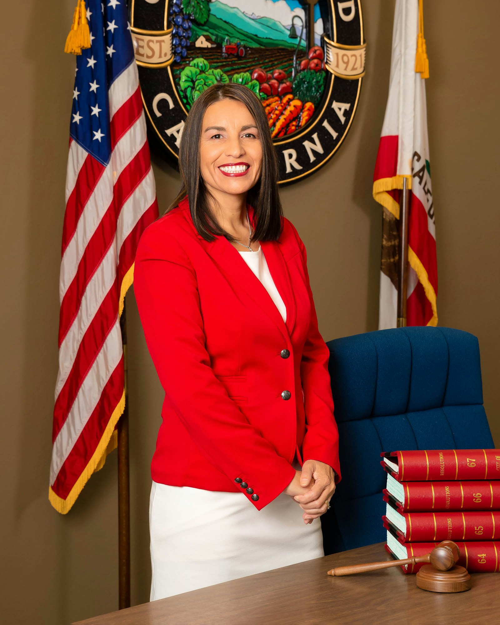 A person in a red suit stands by a desk with books and a gavel, in front of flags and an official seal.