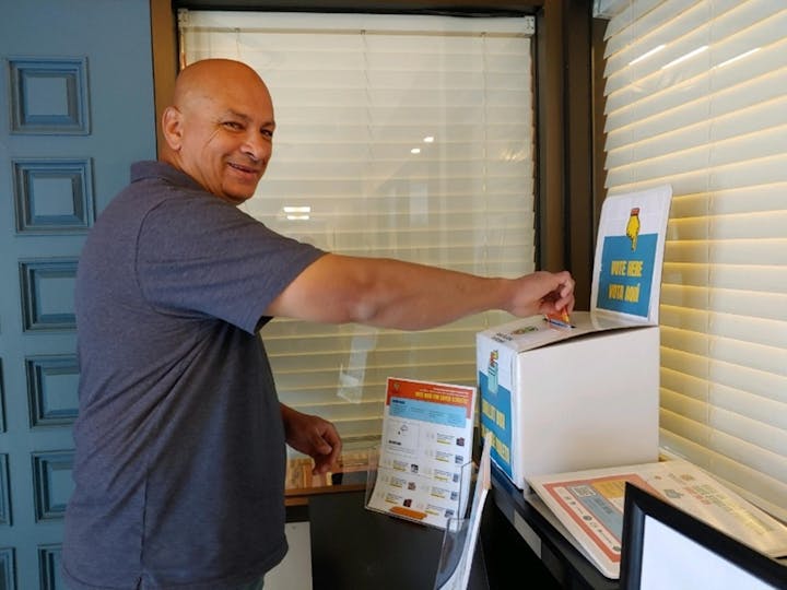 A person is smiling while placing a card into a voting box marked "VOTE HERE".