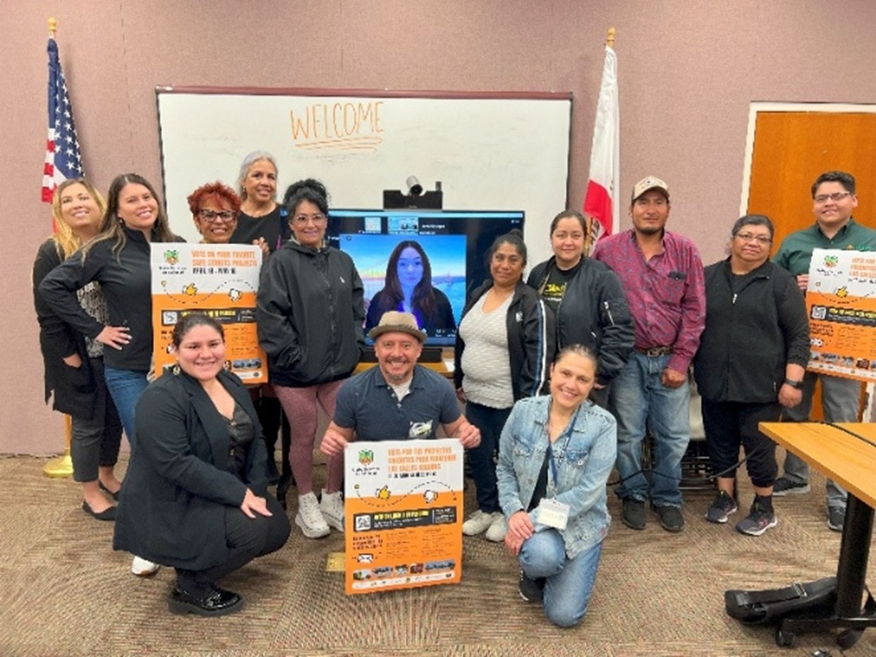 A group of people posing in a room with "WELCOME" on a whiteboard, posters in hand, and a screen showing a video call participant.