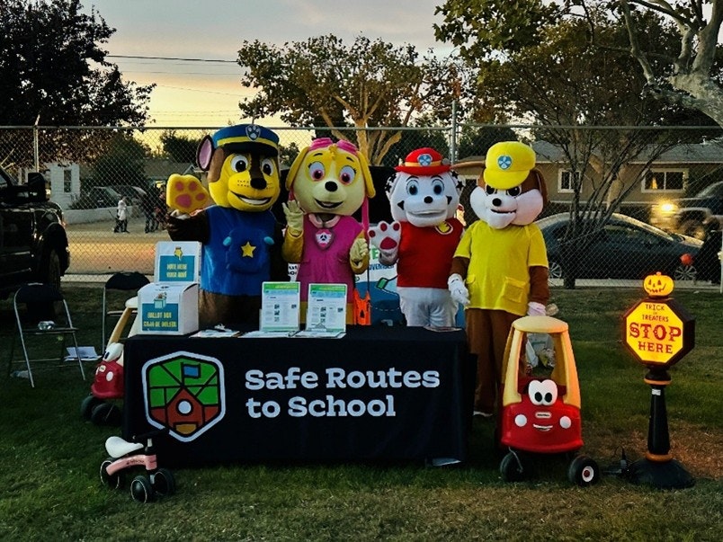Costumed characters at a "Safe Routes to School" booth with signs and a "Trick or Treaters Stop Here" sign.
