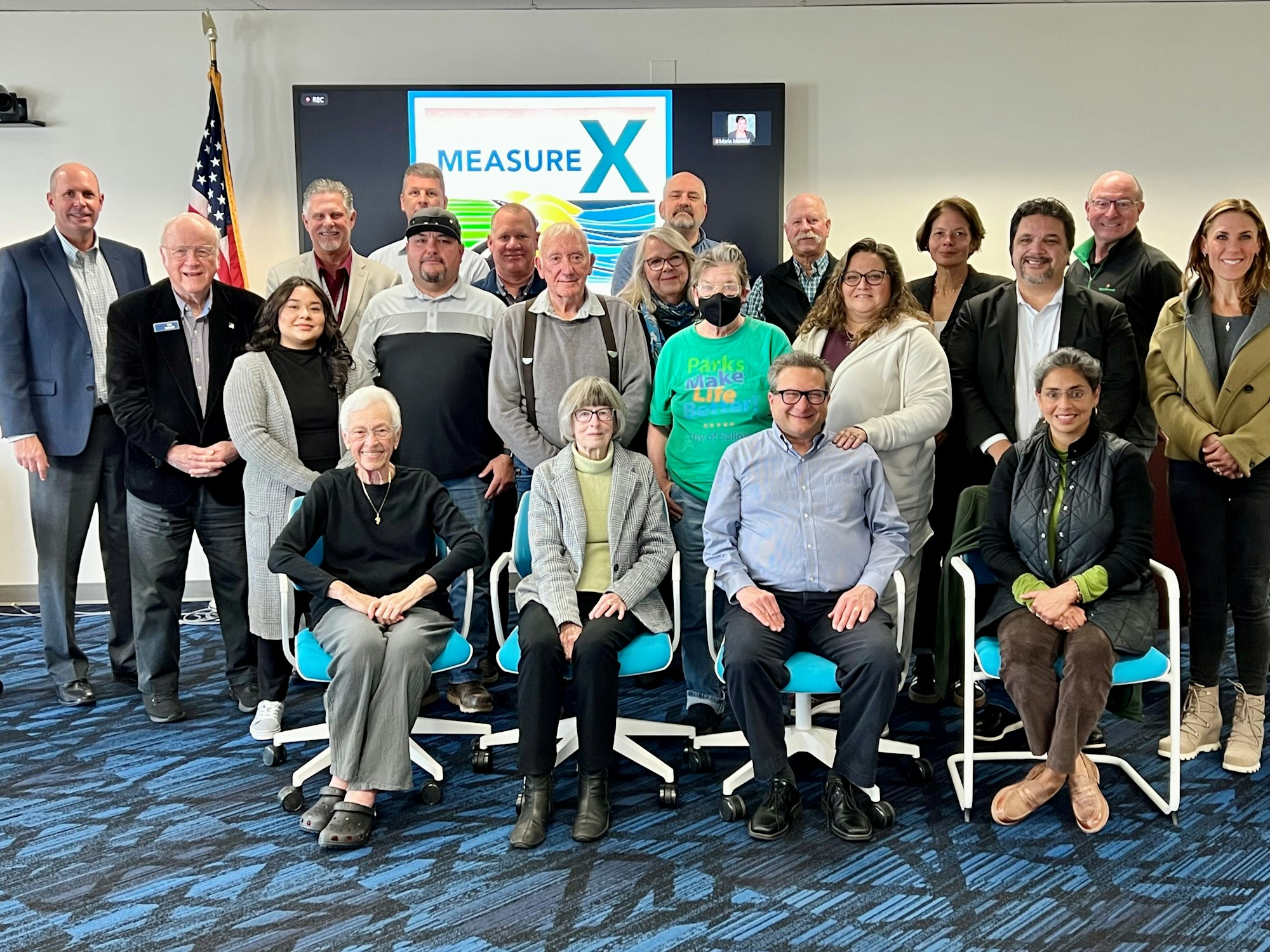 A group of people posing together indoors with a "Measure X" sign in the background.