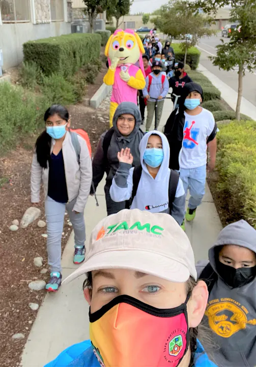 A group of children walking outdoors, wearing masks, with a mascot character in the lead and a person taking a selfie.