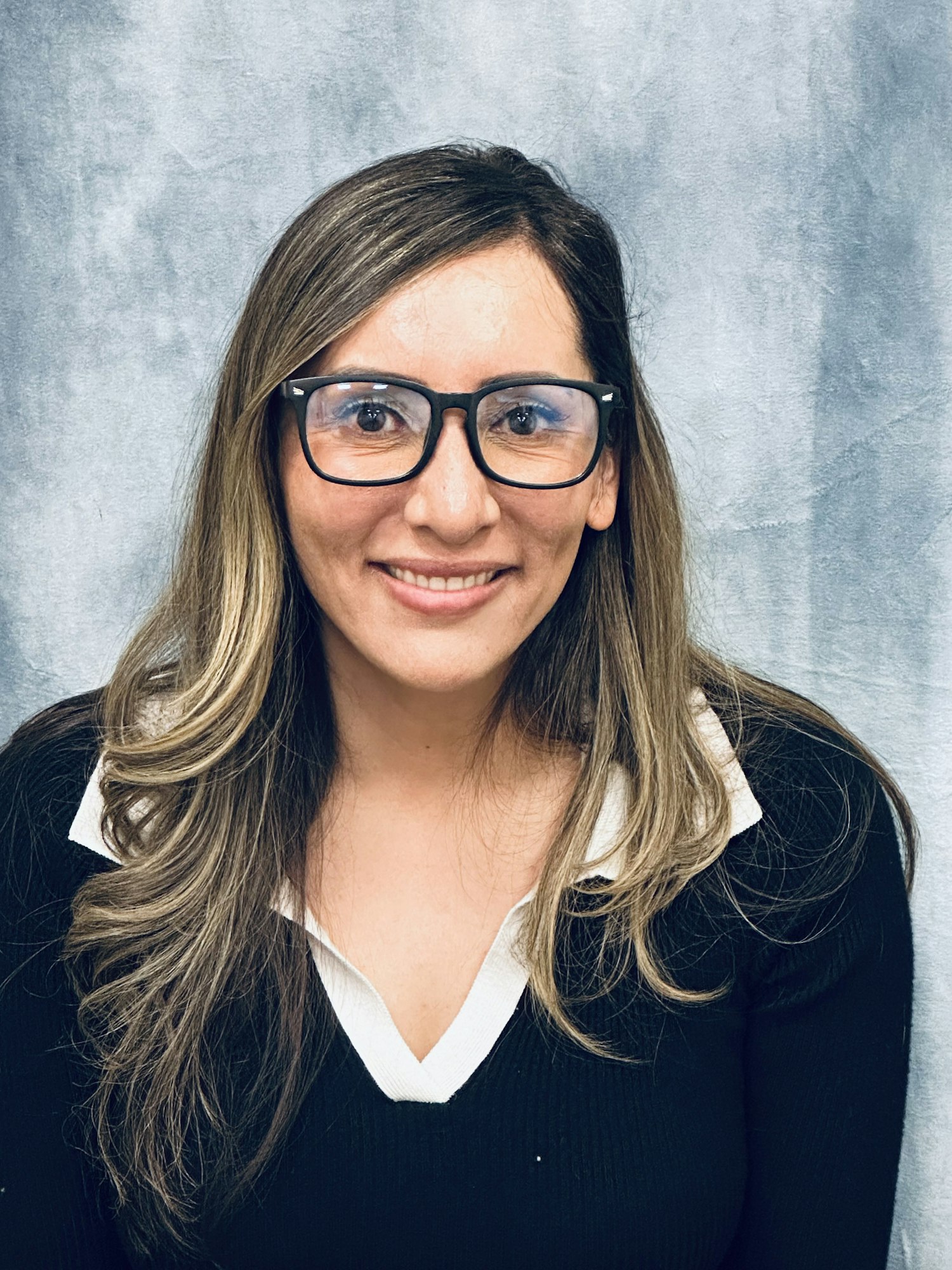 A woman with long hair and glasses smiles against a textured gray background, wearing a black sweater with a white collar.