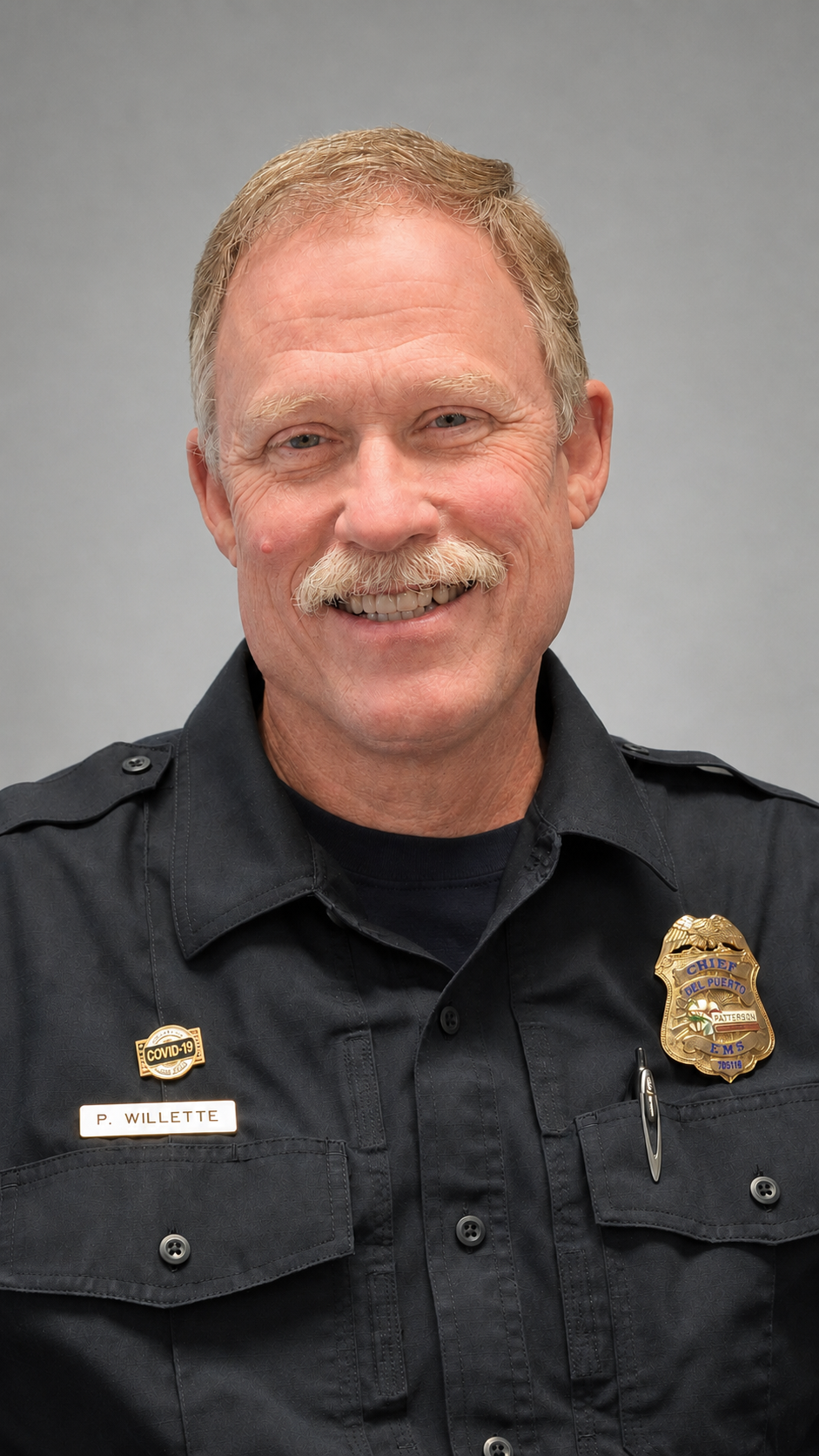 A smiling man in a police uniform with a badge and COVID-19 pin, set against a neutral gray background.