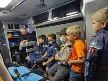 A group of children in scout uniforms sit inside an ambulance with an adult.