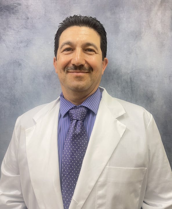 A person in a white coat and a tie, smiling against a gray background.