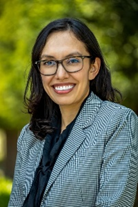 The image shows a smiling individual wearing glasses and a patterned blazer, set against a green background.