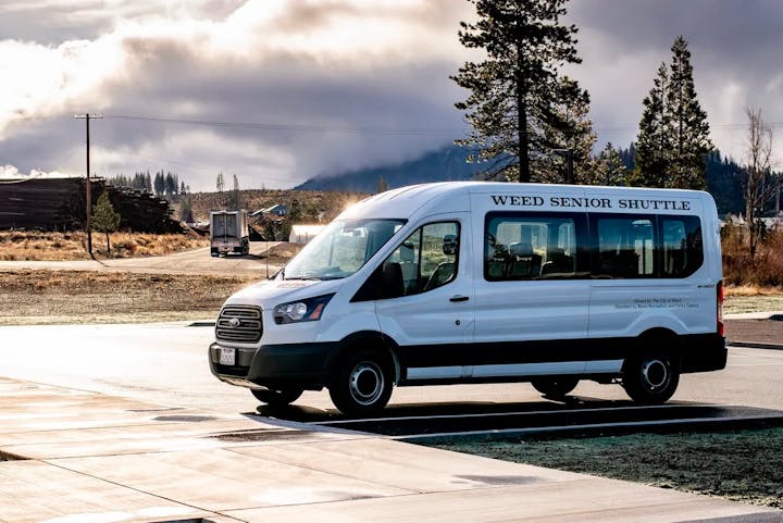 A shuttle bus labeled "Weed Senior Shuttle" parked in an outdoor area with trees and mountains in the background.