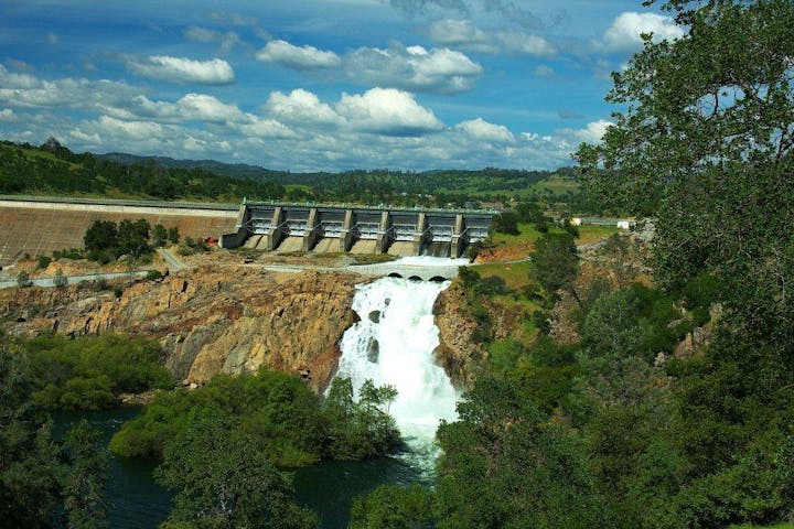 A dam with water flowing down a waterfall, surrounded by trees and hills under a partly cloudy sky.