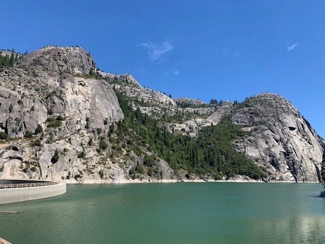 A dam with emerald water and towering rocky cliffs under a clear blue sky.