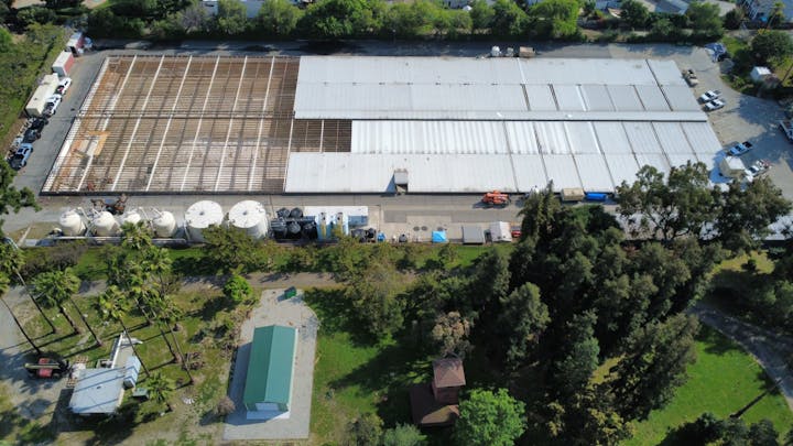 An aerial view of a large industrial building with a partially removed roof, surrounded by trees and equipment.