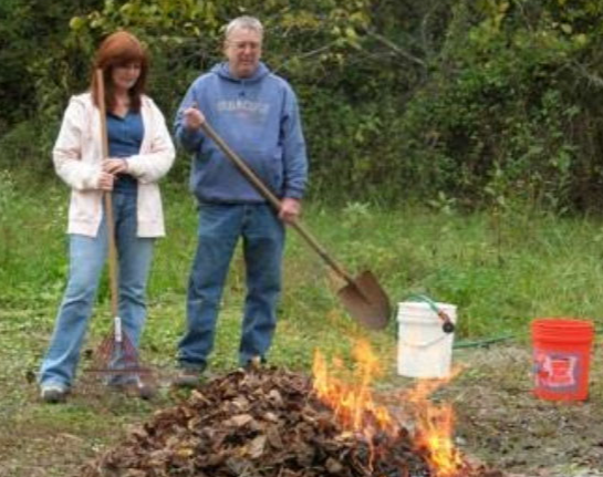 Two people stand by a burning pile of leaves, with rakes and buckets nearby.