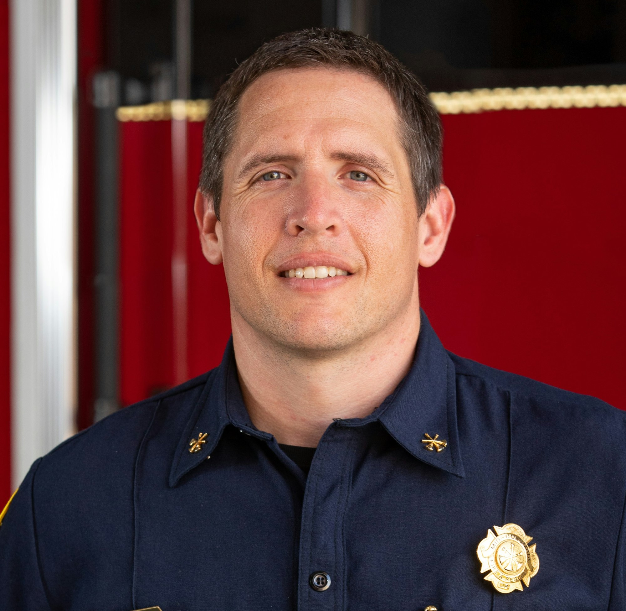 A person in a navy blue uniform with badges, smiling in front of a red background.