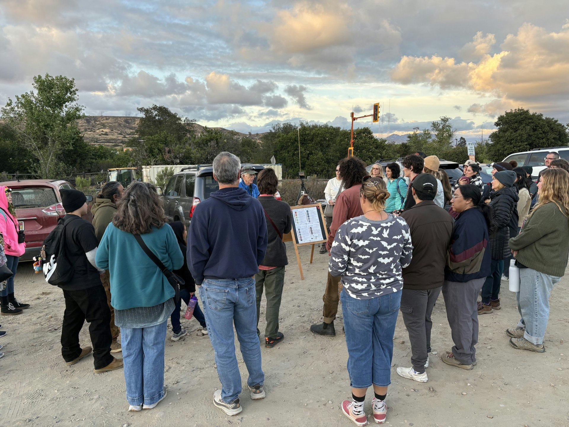 A group of people gathers outdoors, listening to a speaker near parked cars, with a scenic sky and mountains in the background.
