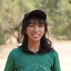 A smiling person wearing a cap, green t-shirt with text, and outdoors.