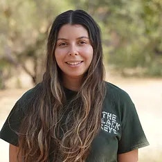 A smiling woman with long wavy hair, wearing a green T-shirt, standing outdoors.
