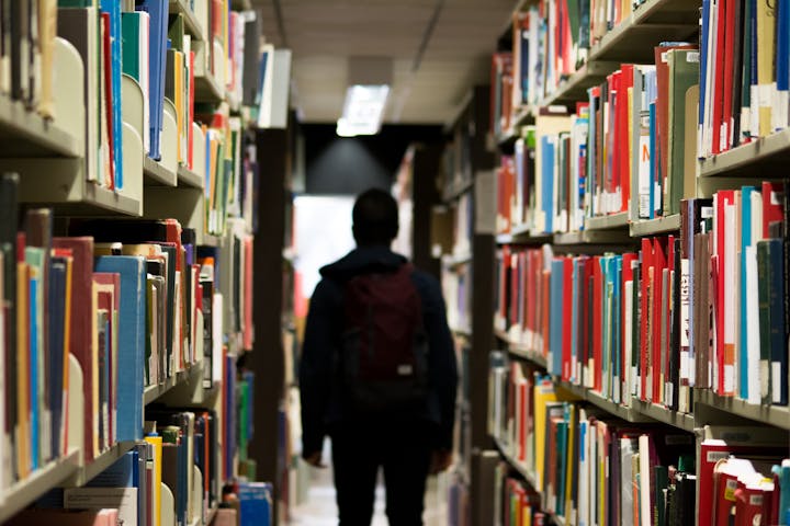 A person walks through a library aisle surrounded by colorful books on shelves. The atmosphere feels quiet and studious.