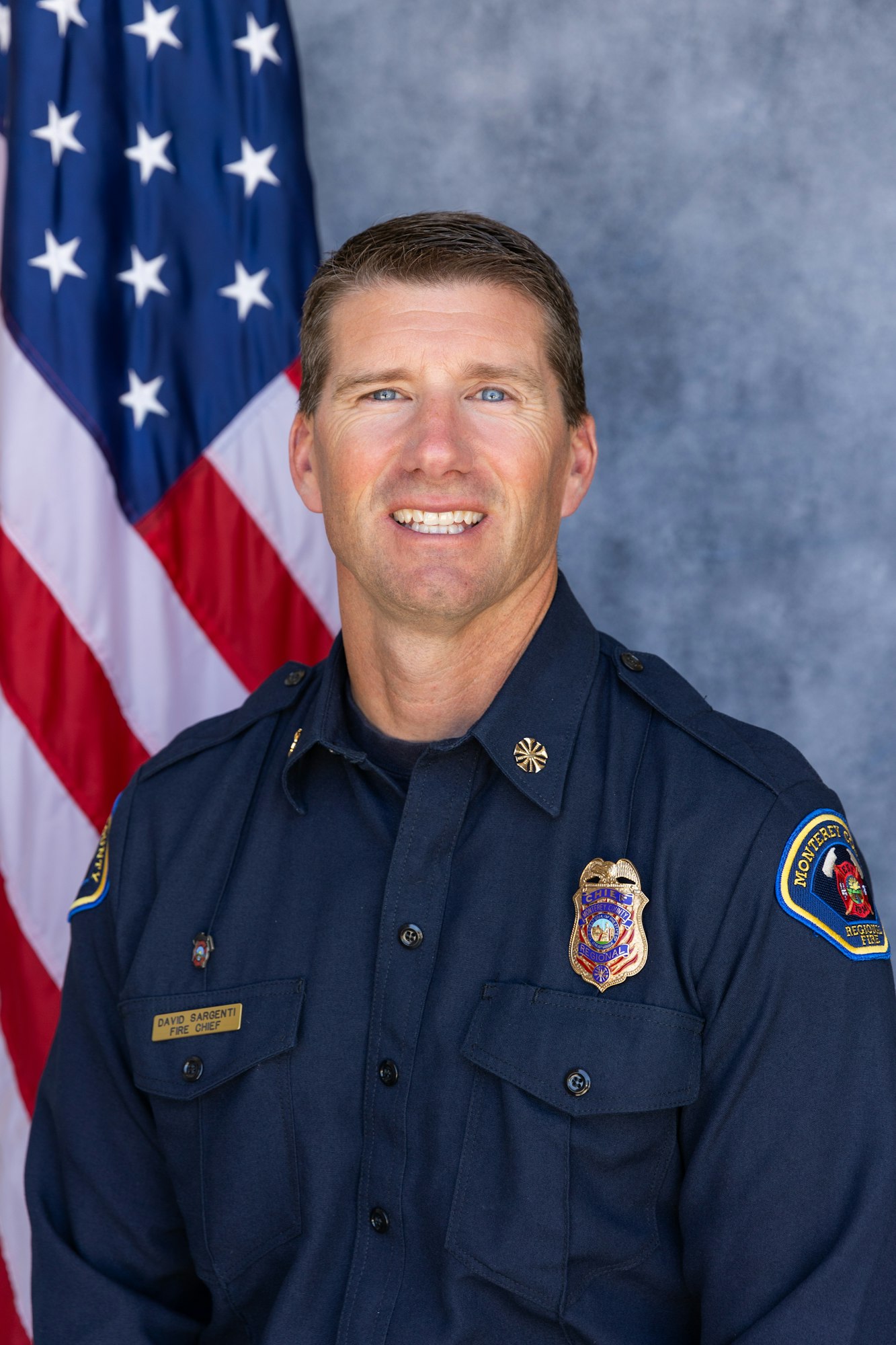 A firefighter in uniform stands in front of a U.S. flag, wearing a badge and name tag.