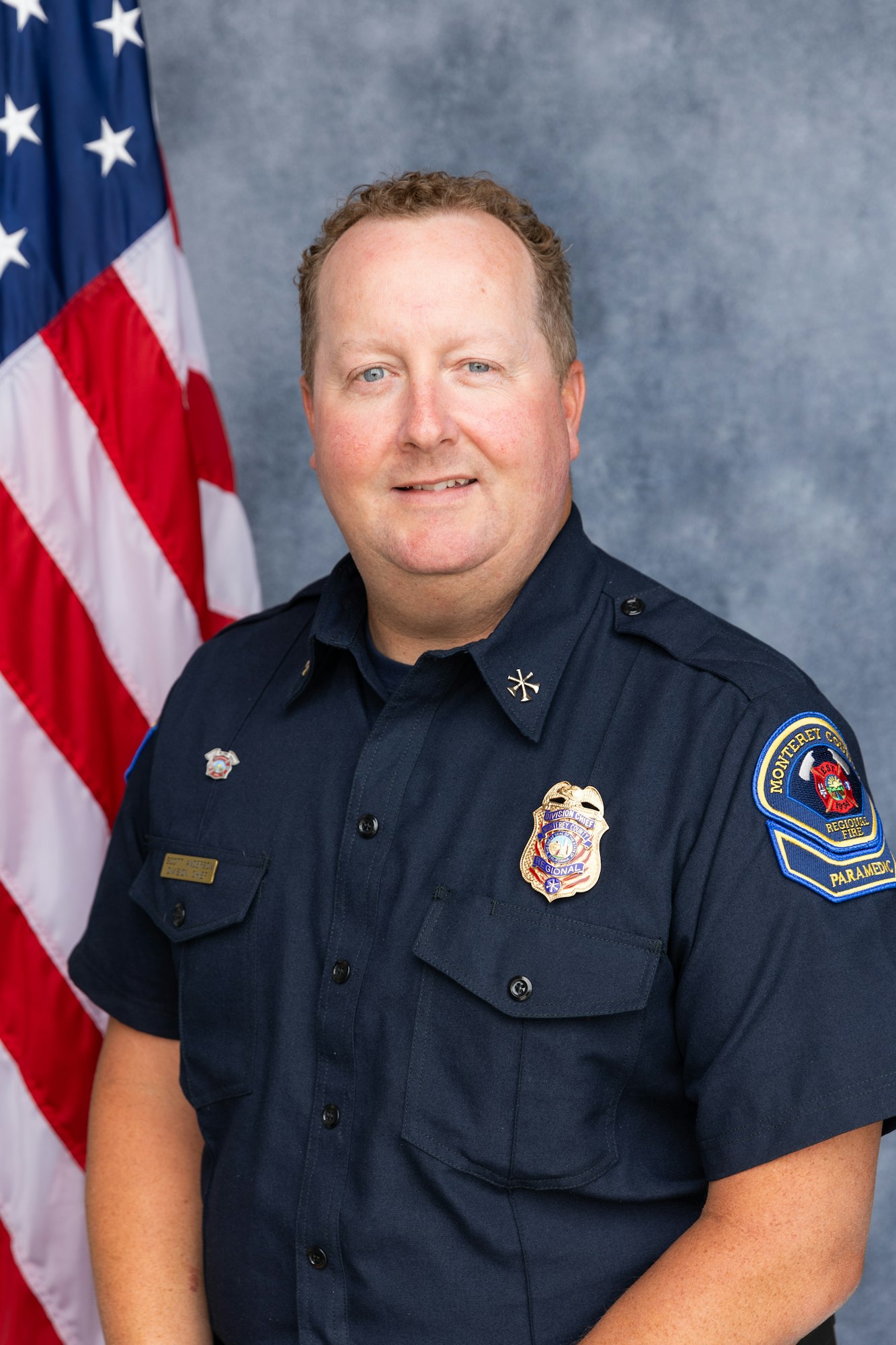 A man in a uniform with a badge and shoulder patch stands in front of an American flag.