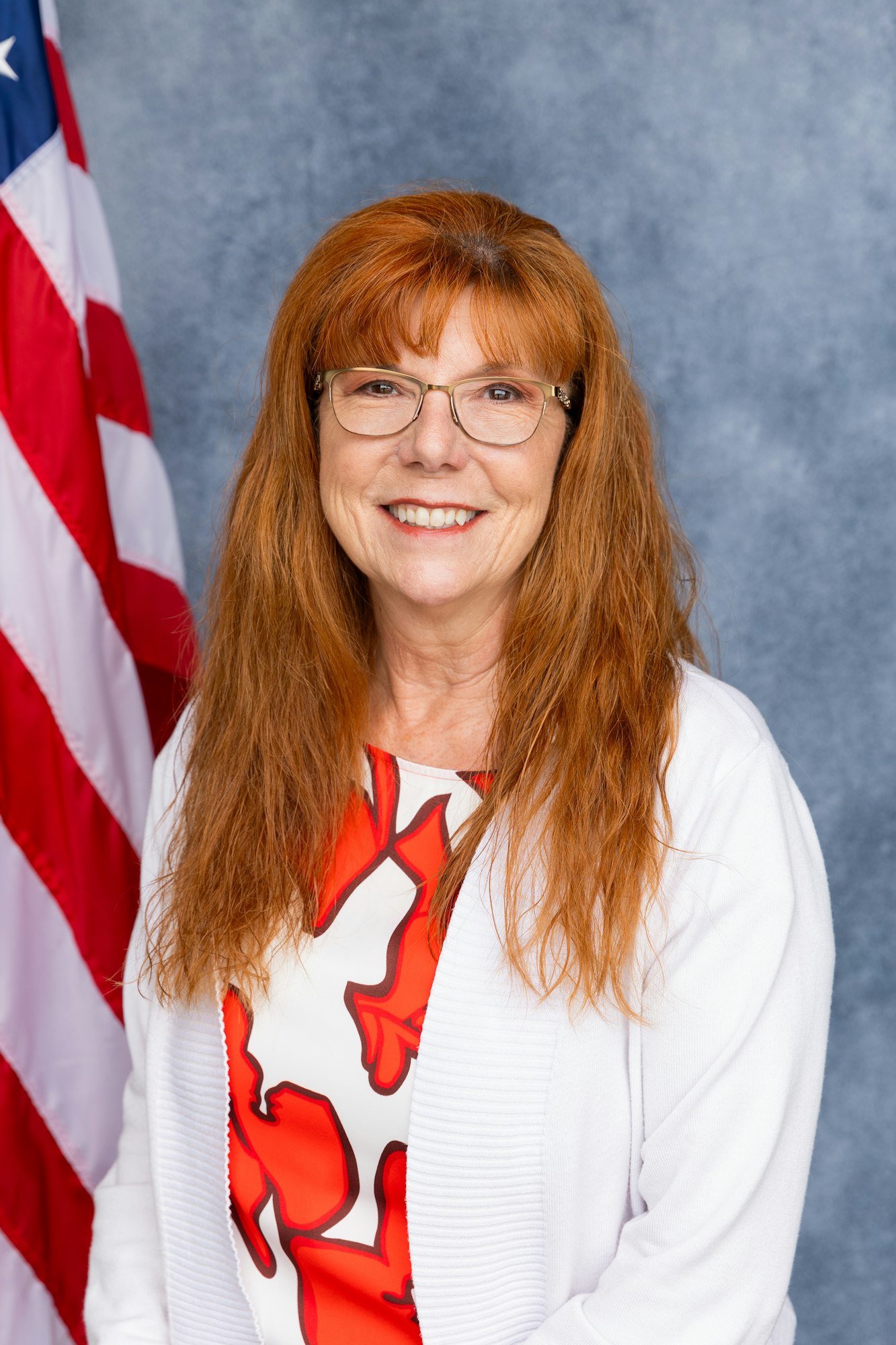 A person with glasses and long red hair smiles in front of a U.S. flag.