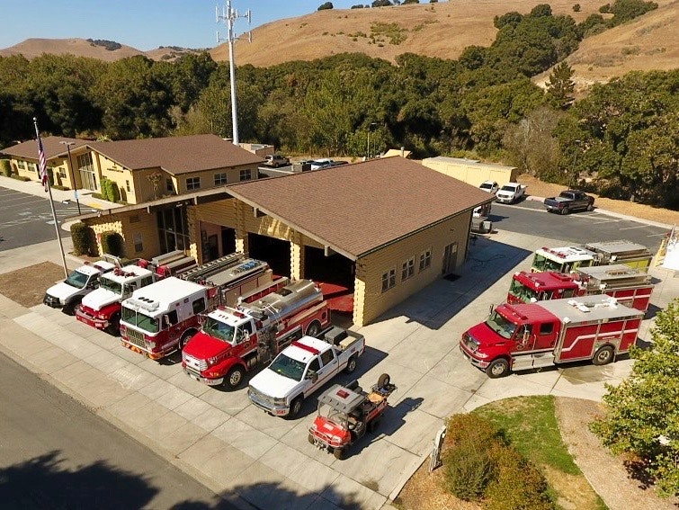 An aerial view of a fire station with multiple fire trucks parked outside, surrounded by greenery and hills.