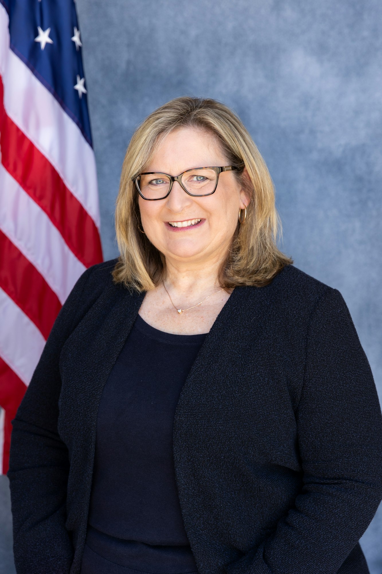 A person smiling, standing in front of a U.S. flag background.