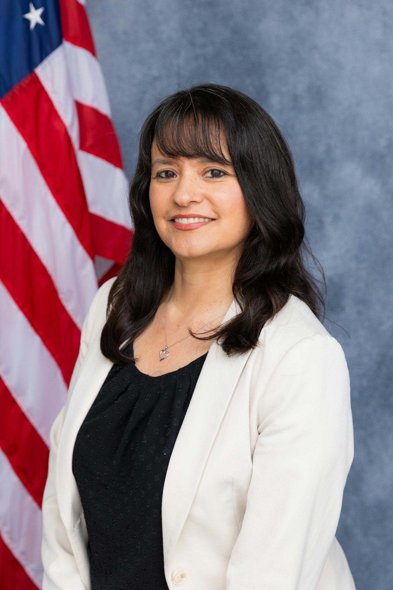 A person in a white jacket and black top poses in front of an American flag.