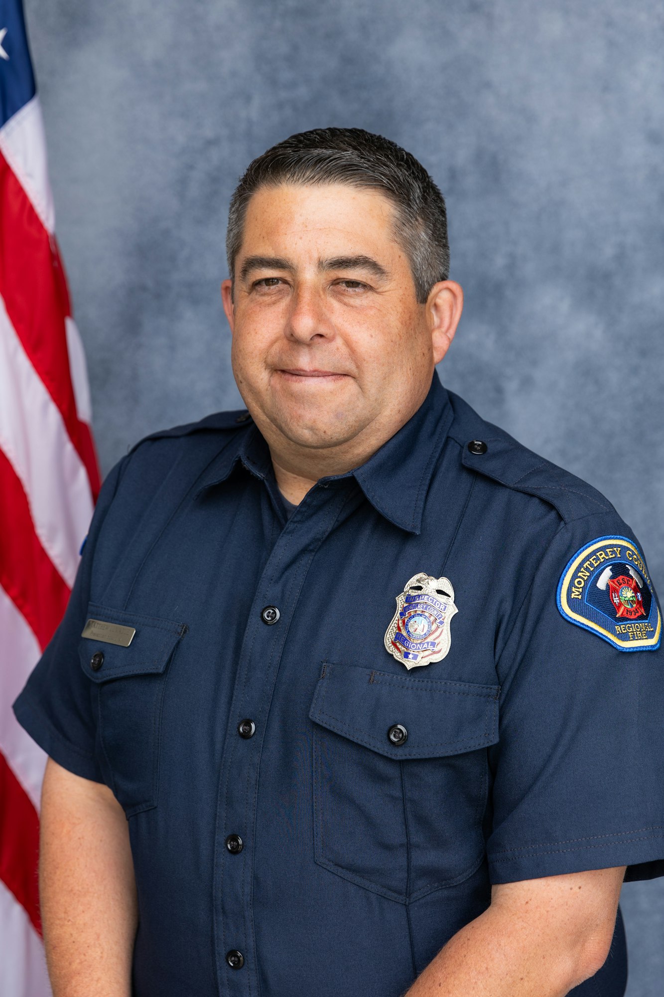 A person in a blue uniform posing with an American flag and a badge labeled "Monterey County Regional Fire."