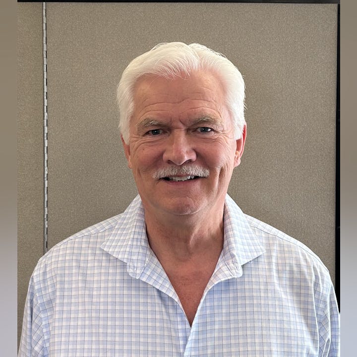 An older man with white hair and a mustache, wearing a checkered shirt, smiling in front of a neutral background.