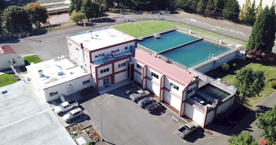 Aerial view of a building with a large outdoor pool, parking lot, and some greenery around.