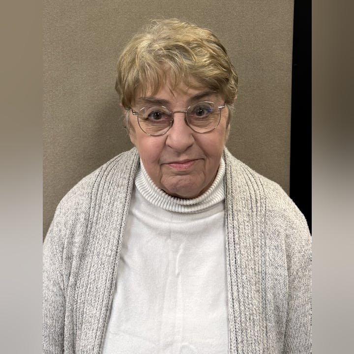 An elderly person wearing glasses, a white turtleneck, and a gray cardigan is looking at the camera.