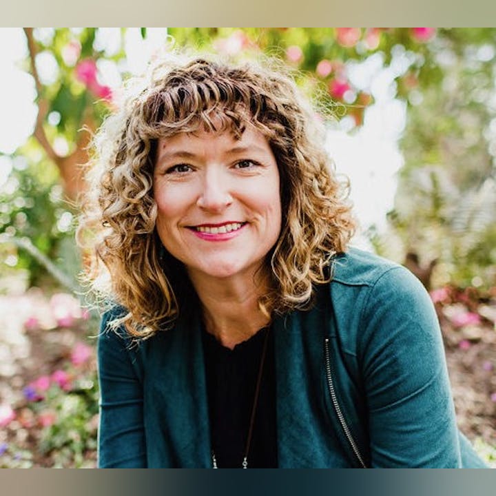 A person with curly hair and a green jacket smiling outdoors with trees and flowers in the background.
