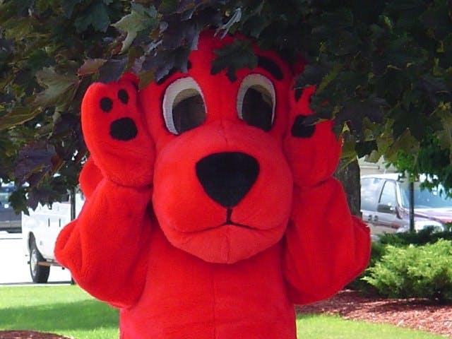 A large red dog mascot costume character under leafy branches in an outdoor setting.