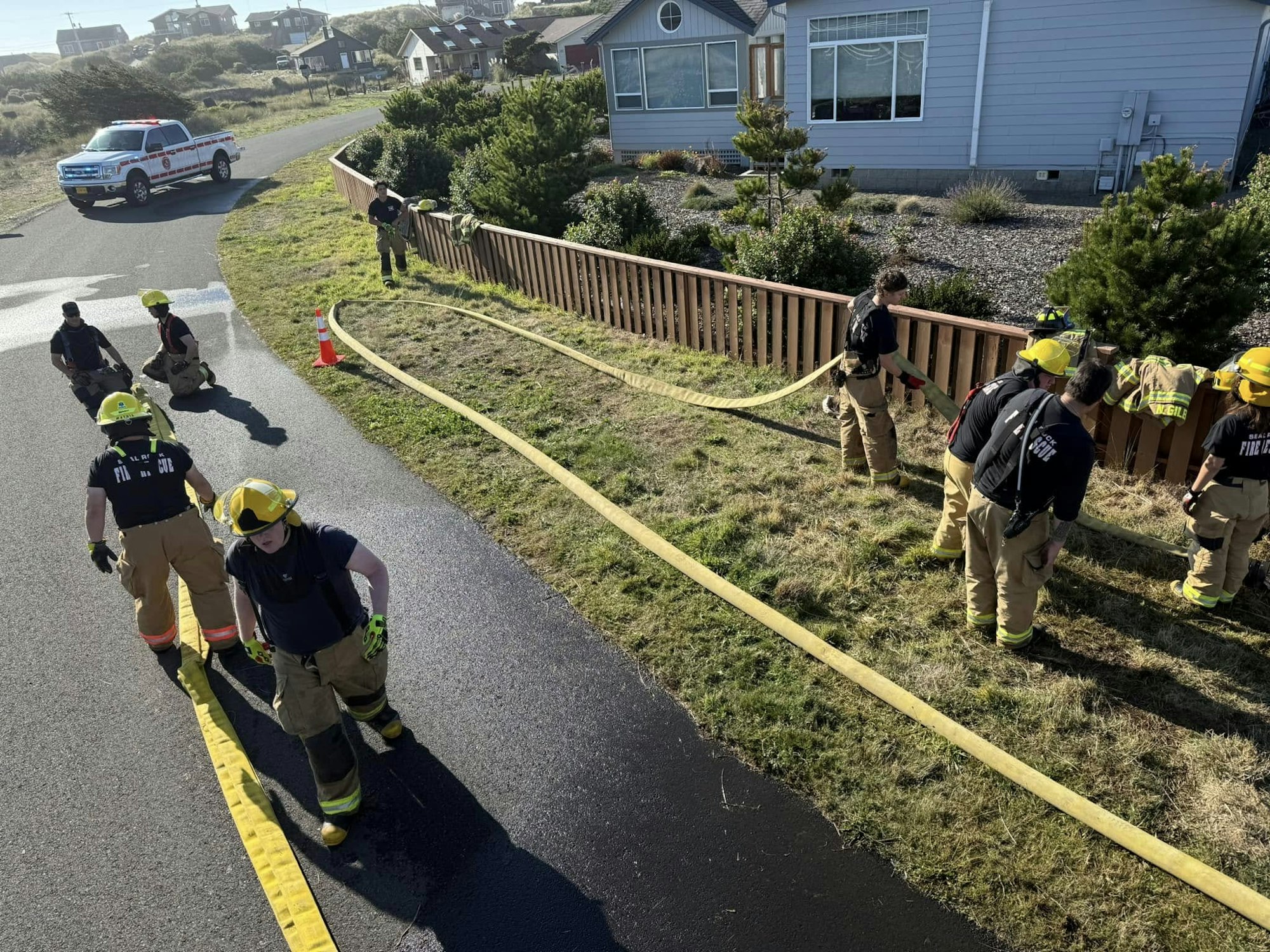 Firefighters laying out hoses next to a house with a fire truck parked nearby on a sunny day.