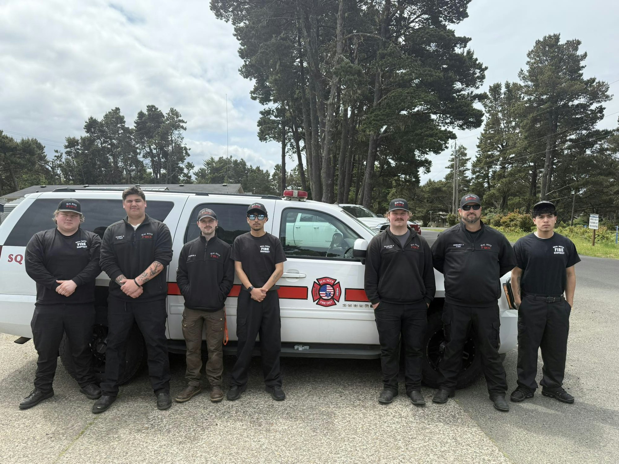 A group of seven people in matching uniforms stand in front of a fire department vehicle, surrounded by trees.