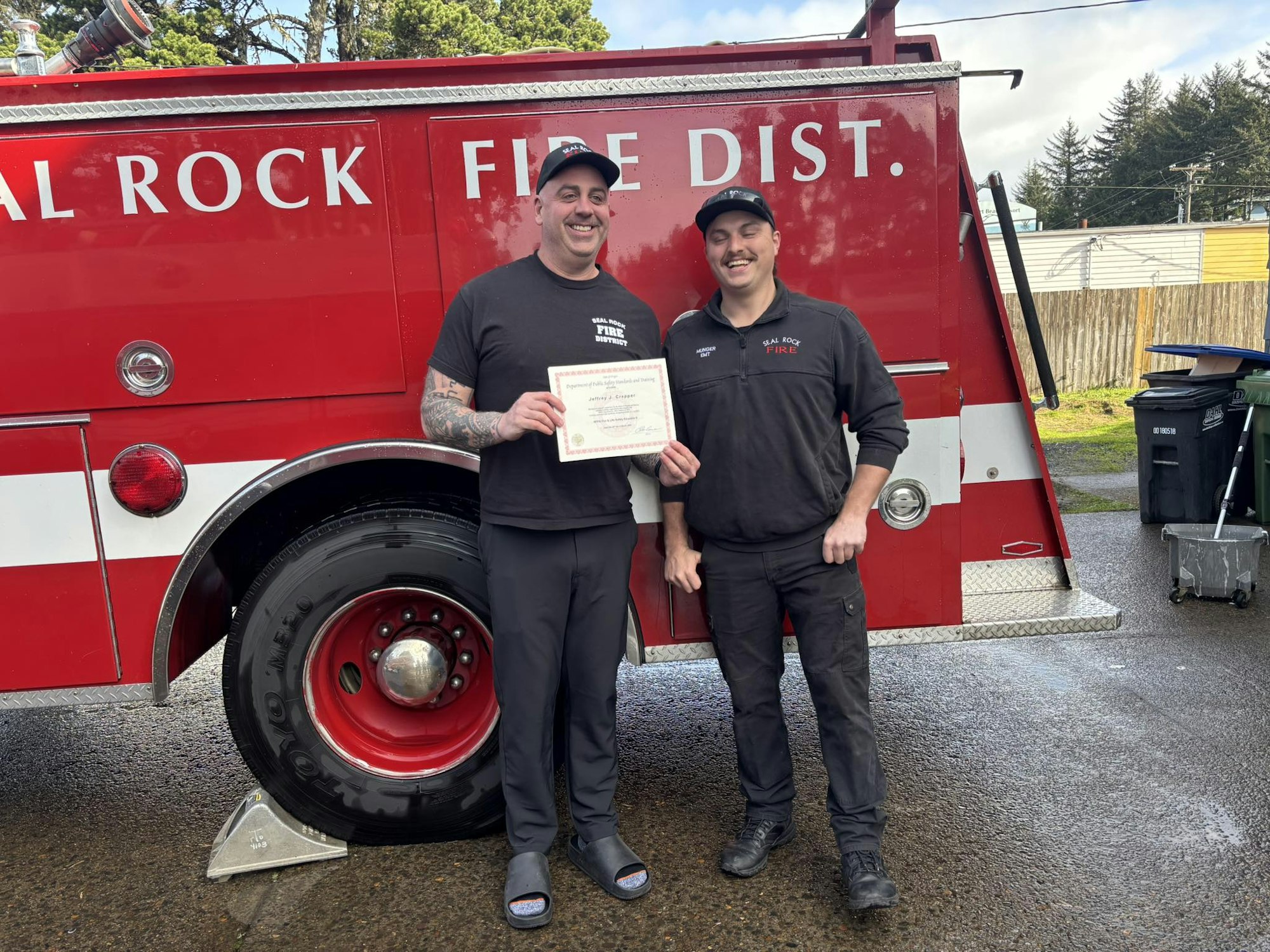 Two people stand by a fire truck, one holding a certificate, both smiling.