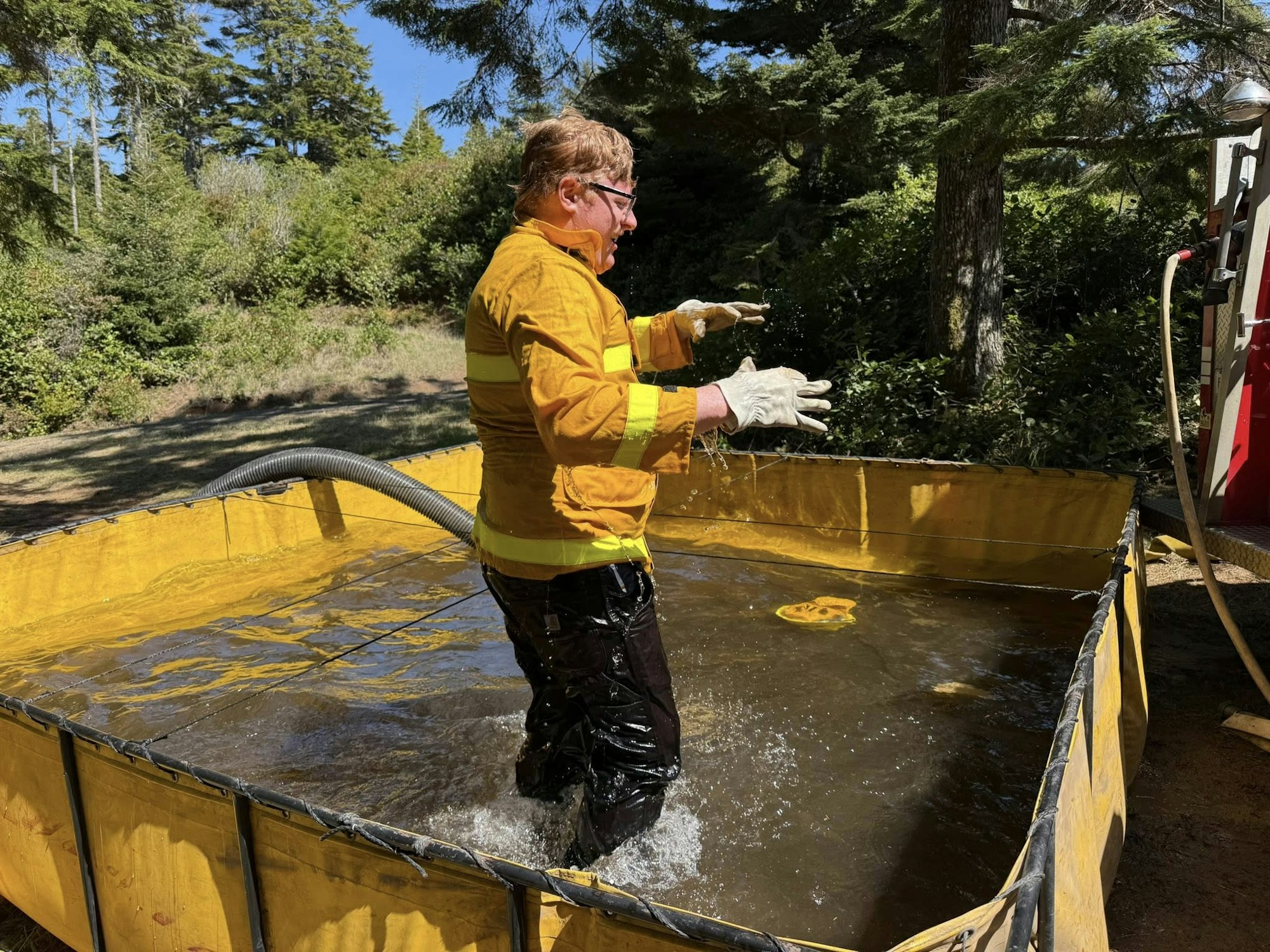 A person in yellow protective gear stands in a portable water tank outdoors, with a hose connected to it.
