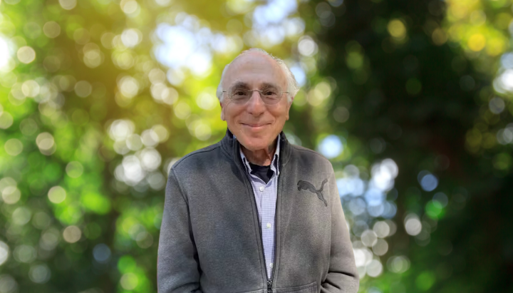 A smiling person in a gray jacket stands outdoors with a bokeh background of leaves.