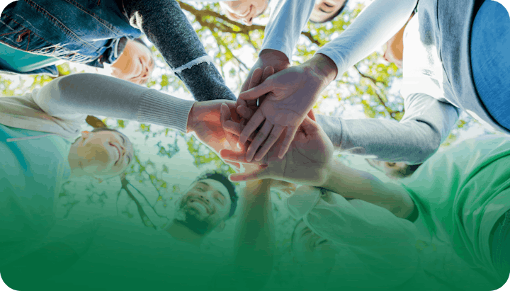 A group of people joining hands in a circle, viewed from below with trees in the background.