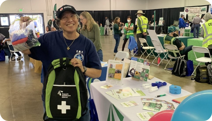 An EMT in a blue shirt smiles, holding a first aid bag at a community event, with informational tables in the background.