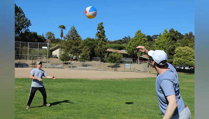 Two people are playing catch with a beach ball on a sunny day in a park. Trees and a sports field are visible in the background.