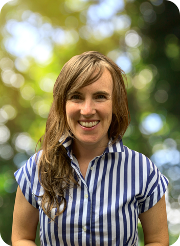 A smiling person with wavy hair, wearing a striped shirt, is outdoors with a blurred green background.