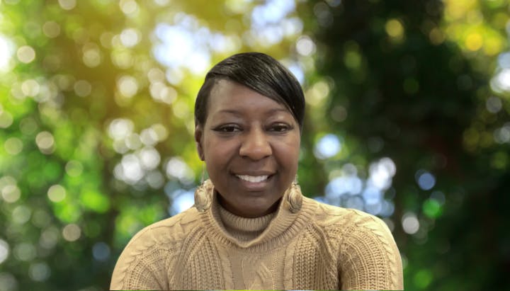 A person in a sweater is smiling with a blurry background of trees and sunlight.